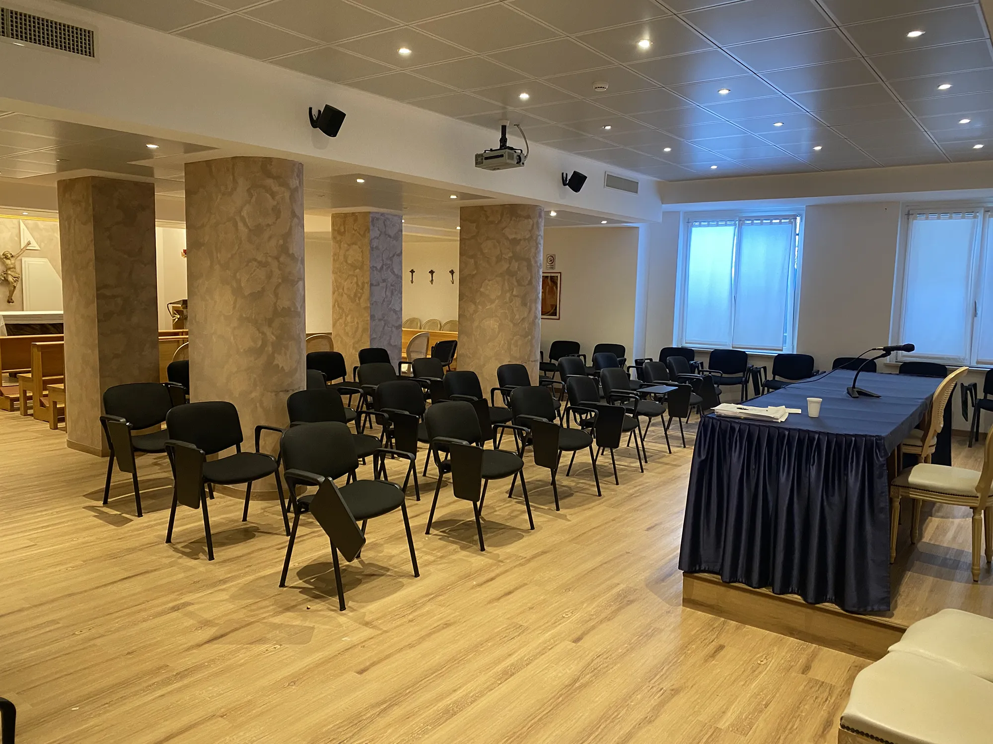Nell'immagine la sala meeting dell'hotel con ordinate file di sedie nere. Al centro ci sono 2 colonne marrone chiaro. Di fronte alle sedie c'è il tavolo dei relatori coperto con una tovaglia blu.