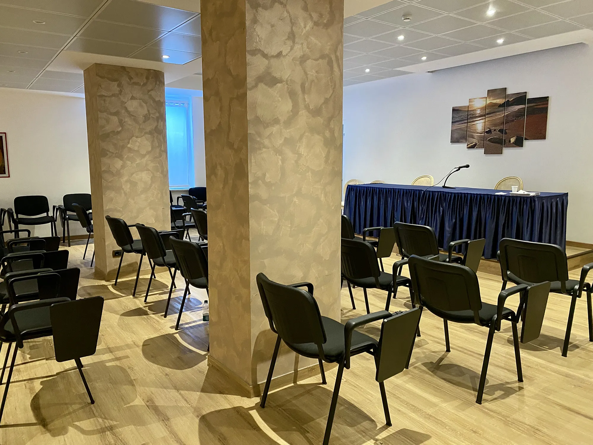 Nell'immagine la sala meeting dell'hotel con ordinate file di sedie nere. Al centro ci sono 2 colonne marrone chiaro. Di fronte alle sedie c'è il tavolo dei relatori coperto con una tovaglia blu.