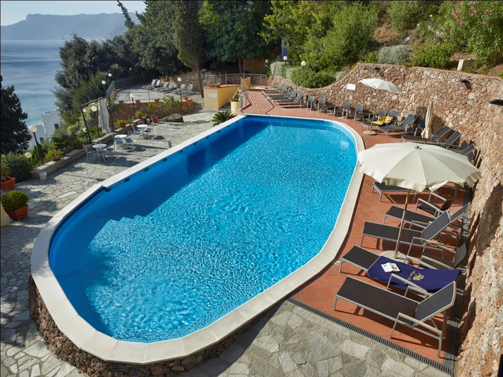 Nell'immagine la piscina e l'area solarium dell'hotel. Al centro c'è la piscina e sul lato destro lettini e sedie per prendere il sole. Ci sono anche alcuni ombrelloni, sia chiusi che aperti. A sinistra della piscina ci sono alcuni tavolini con sedie. Intorno ci sono degli alberi e a sinistra c'è uno scorcio di mare.