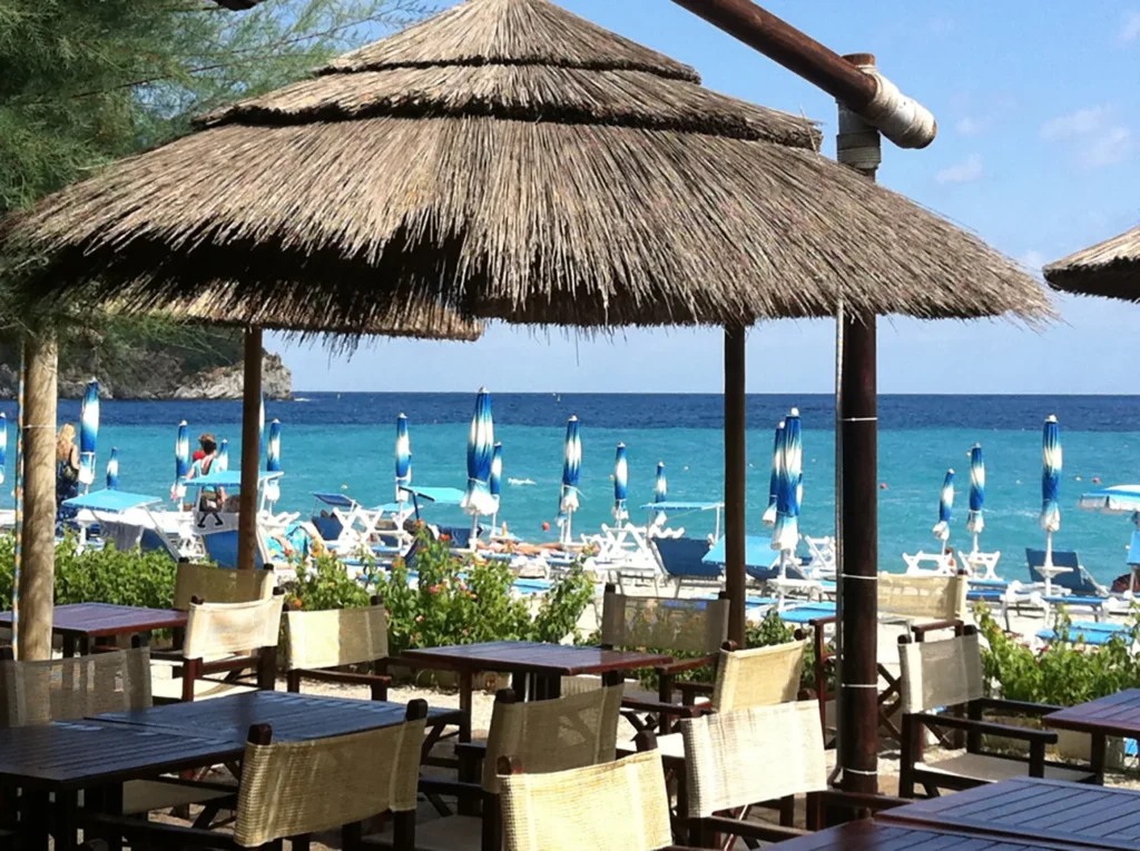 Nell'immagine in primo piano i tavoli e le sedie del ristorante dell'hotel che si trova sula spiaggia. Un grande ombrellone di paglia copre i tavoli dal sole. Dietro si vedono gli ombrelloni e i lettini della spiaggia e il mare.