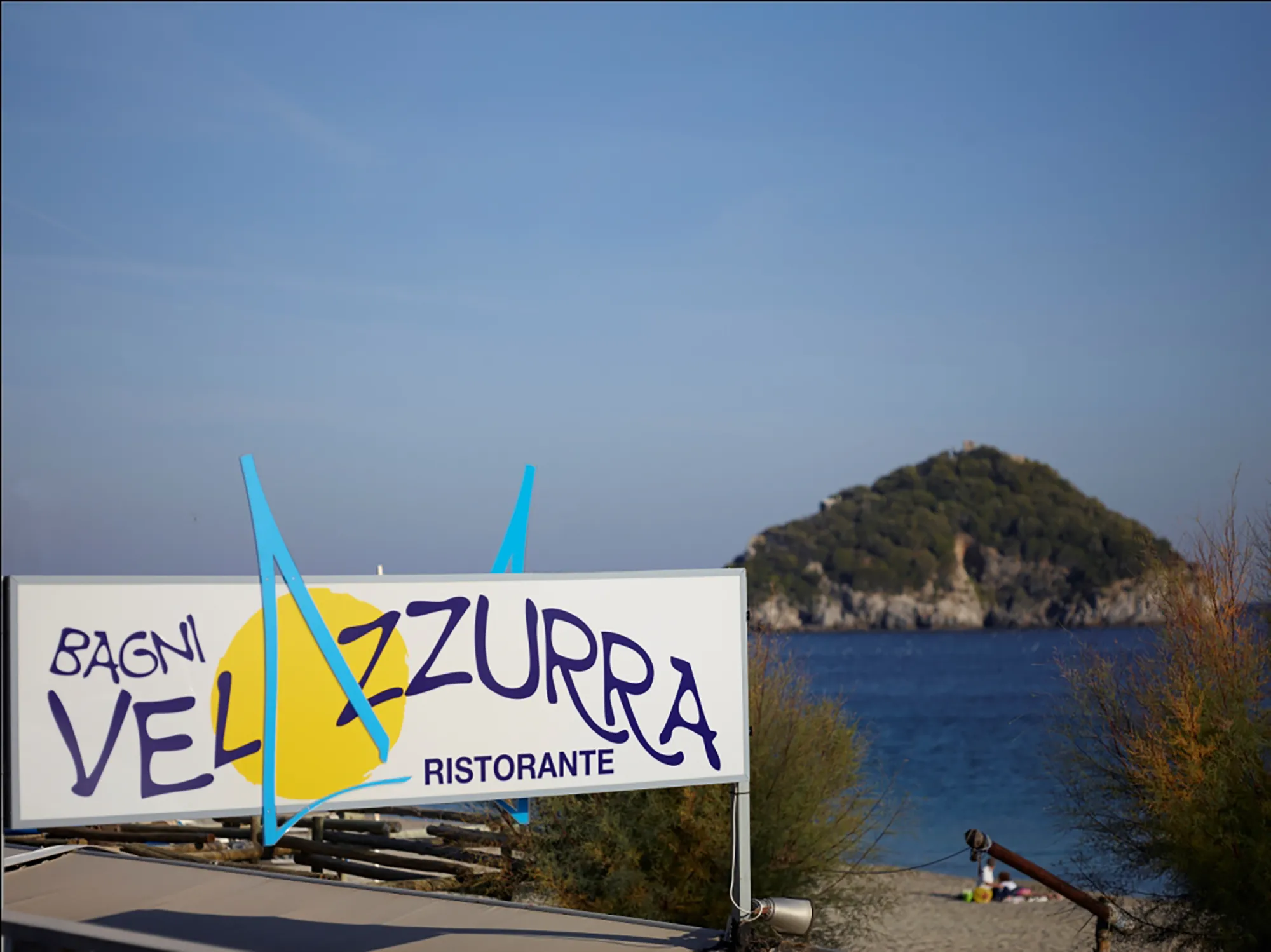 Nell'immagine c'è l'insegna dei bagni Velazzurra, la spiaggia dell'hotel. Dietro si vede la spiaggia, il mare e l'isola di Bargeggi
