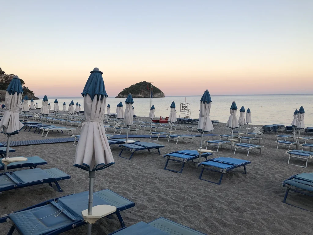 Nell'immagine la spiaggia convenzionata dell'hotel con file ordinate di lettini azzurri e ombrelloni bianchi chiusi. Di fronte si vede l'isola di Bergeggi.