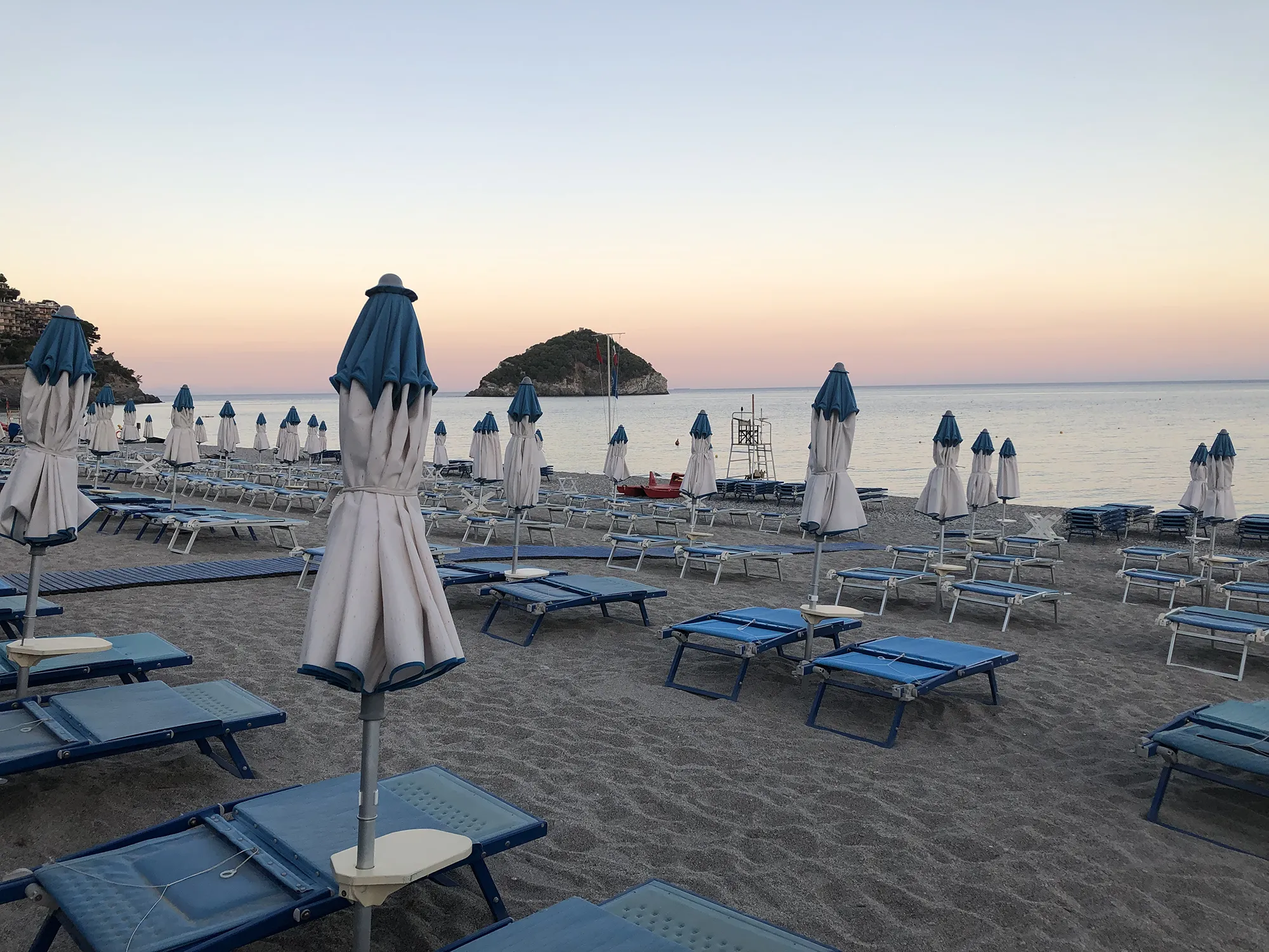 In the picture the hotel's private beach with neat rows of blue loungers and closed white umbrellas. In front you can see the island of Bergeggi.